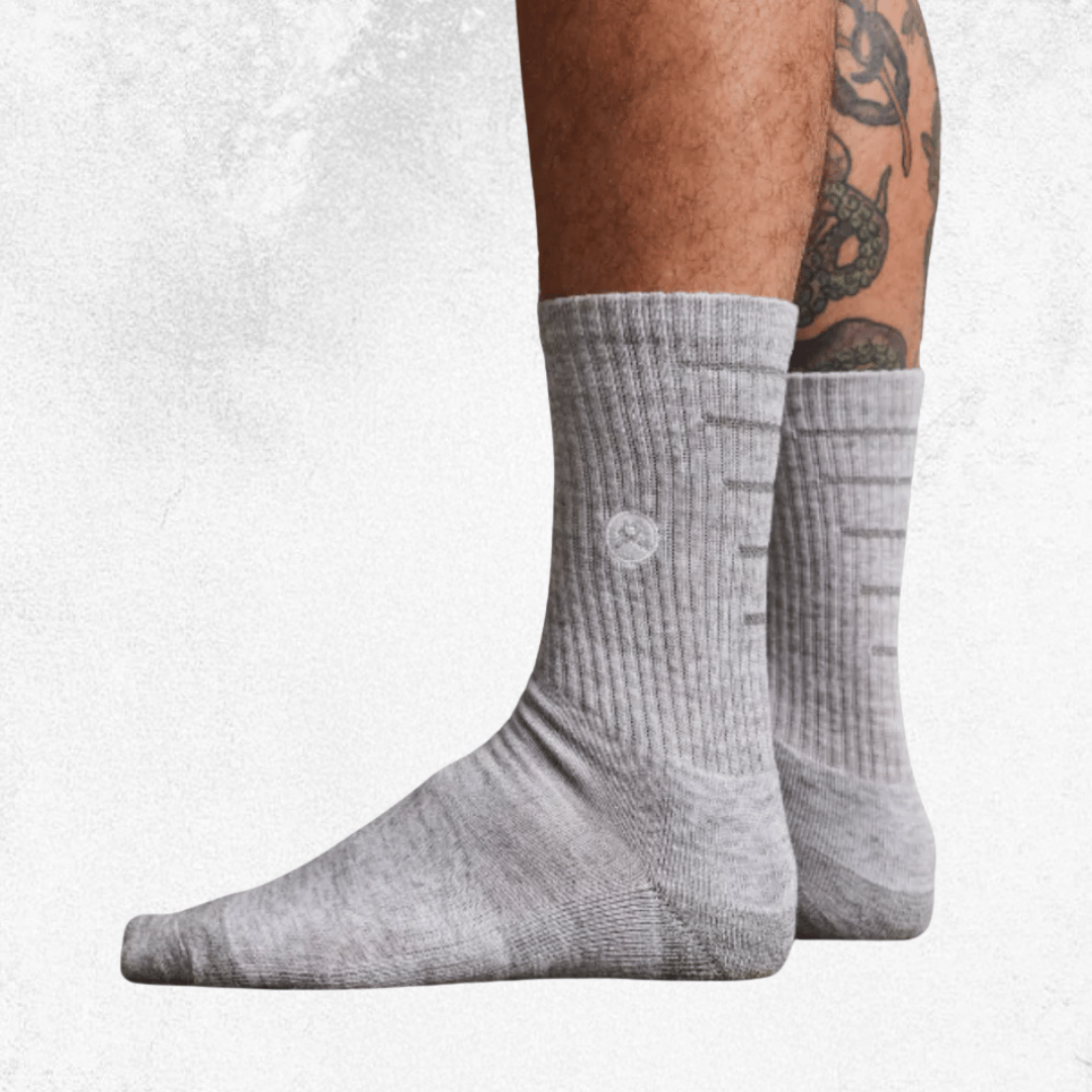 A pair of light gray ribbed crew socks worn on a person's feet, shown against a light textured gray background. The person's lower legs are visible, with one leg featuring a black snake tattoo.