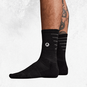 Black crew socks with a small white circular logo on the side, shown worn on a person's feet against a light textured background.