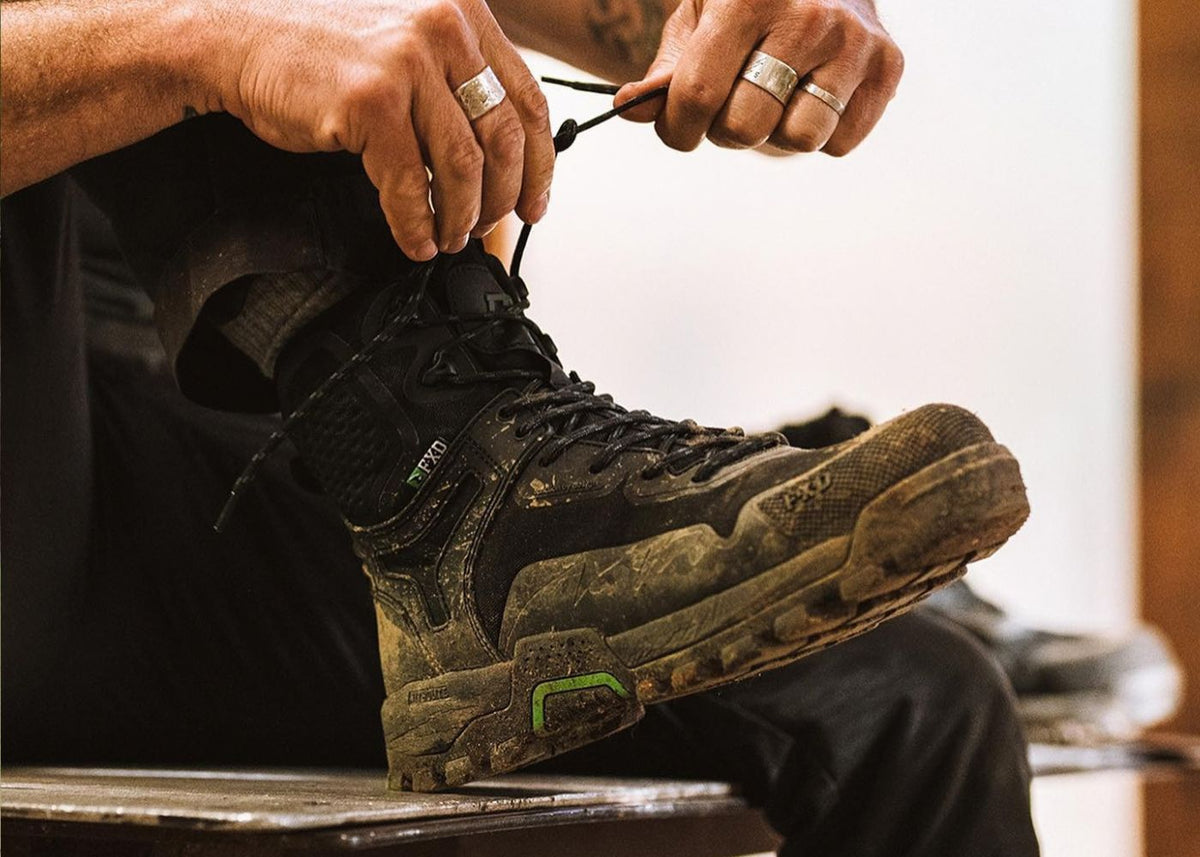 A close-up lifestyle shot of a person tying the laces of a rugged, muddy black FXD Work boot. The person's hands and lower legs are visible, along with silver rings on their fingers.
