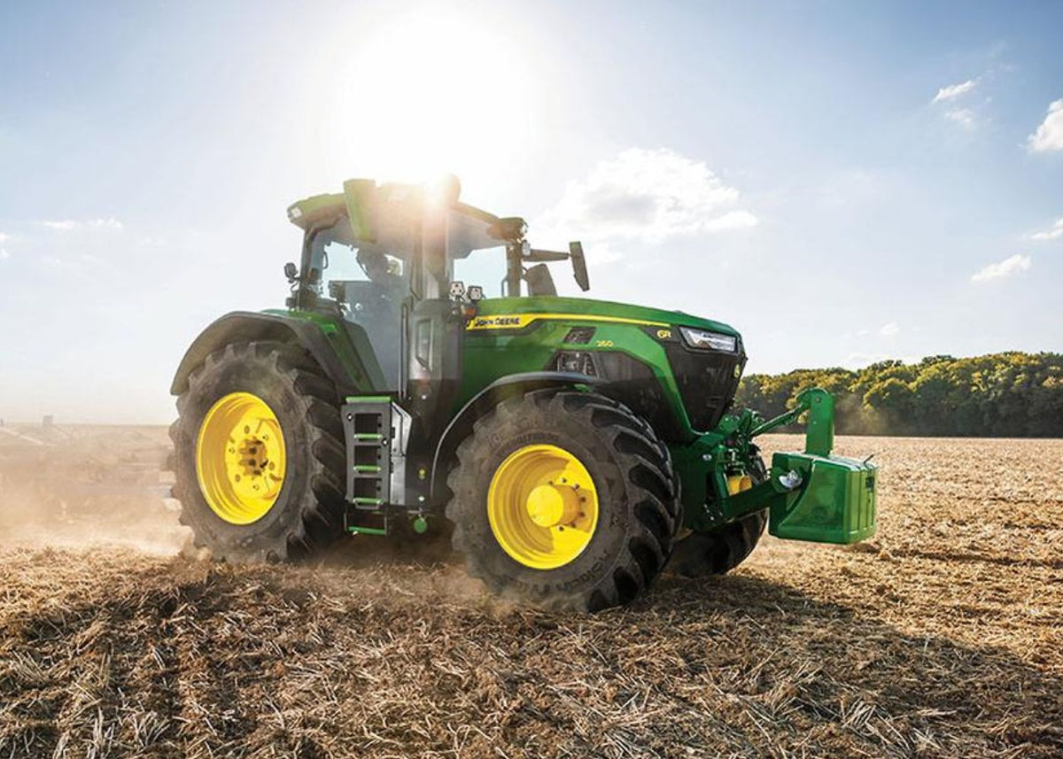 A green John Deere tractor with yellow wheels is shown in a real outdoor field under bright sunlight, pulling or working the soil. Because a real environment is present, this is Lifestyle rather than Product/Packaging.