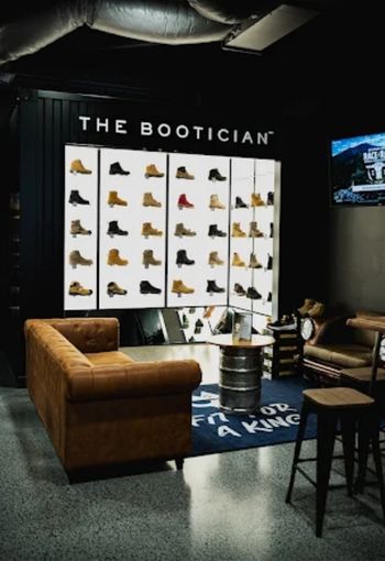 A retail display for The Bootician boots, featuring a large illuminated wall display of various ankle boots, a brown leather Chesterfield sofa, a metal barrel coffee table, and a blue branded rug on a dark industrial floor.