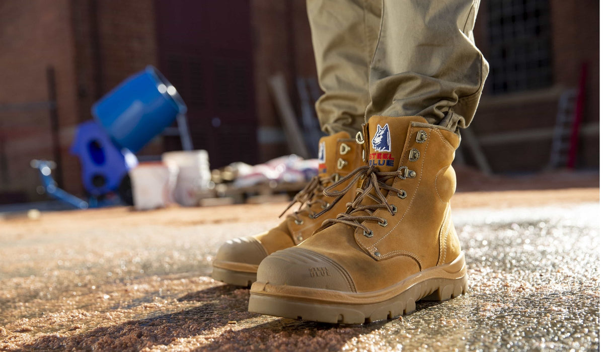 A close-up of a person wearing wheat coloured steel blue work boots with khaki pants, standing outdoors on a concrete surface with a blurred brick wall and blue equipment in the background.