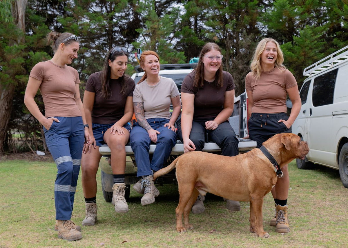 Five women in casual outdoor work clothes and a tan dog stand in front of a white van on a grassy area with trees in the background. They are all wearing Zadie Workwear clothing.