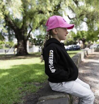 Kid sitting in park with pink hat on and wearing Kids Cat Workwear Trademark Banner Zip Sweatshirt