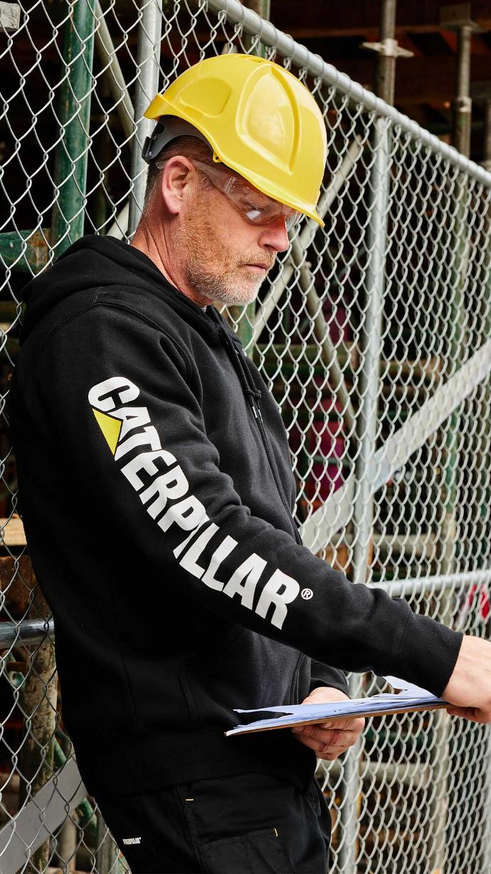 Man wearing CAT Midweight Banner Full Zip Hoodie in colour black while reading from a clipboard on a construction site