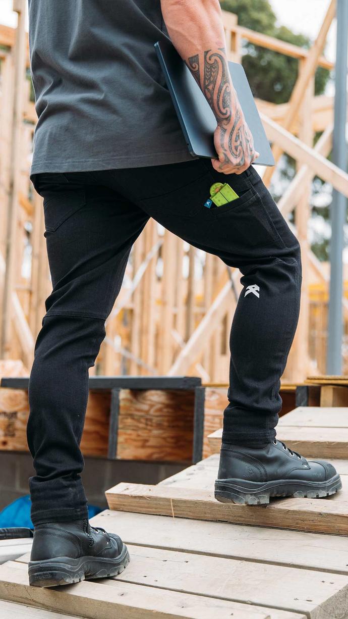 Tradie with tattoos wearing black Anthem Tech Stretch Denim Jeans on construction site.